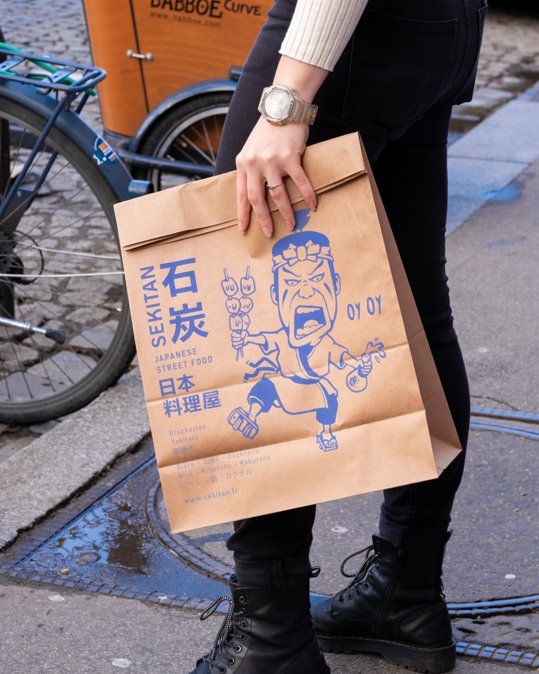 Person carrying a block bottom paper bag with Sekitan Japanese street food branding in blue print, walking on a city street with a bicycle in the background.